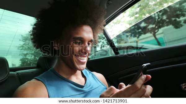 Black man in backseat of car typing\
on cellphone smiling, riding taxi while on\
smartphone