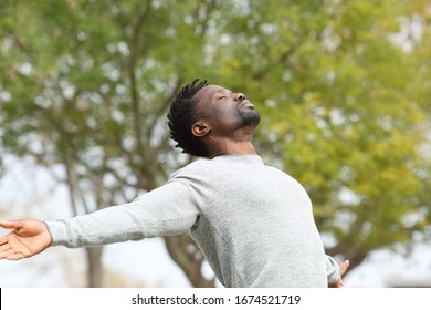 Black man breathing fresh air stretching arms in a park with a green tree in the background