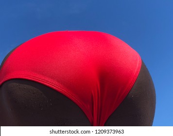 A Black Man In A Bright Red Racing Bathing Suit Bends Over Against A Blue Sky At The Beach.