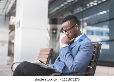 black man businessman in a business suit and glasses sits on a bench and reads a newspaper against the backdrop of a modern city - Shutterstock ID 1151873276