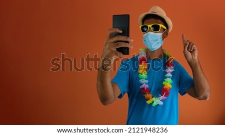 Black man in carnival costume and pandemic mask holding  a mobile phone isolated on orange background. 