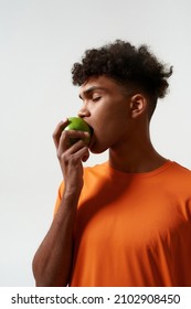 Black Man With Closed Eyes Eating Green Tasty Organic Apple. Concept Of Dieting And Healthy Eating. Young Curly Sporty Guy Wearing Orange T-shirt. Isolated On White Background. Studio Shoot