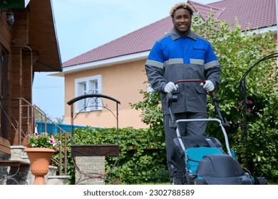 Black man in coveralls uses a lawn mowing machine in the backyard. A smiling African man in overalls with a lawn mower mows the green grass in a modern garden. Professional lawn care service. - Shutterstock ID 2302788589
