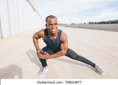 Black man doing stretching before running in urban background. Young male exercising listening to music with headphones.