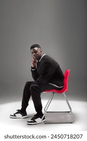 A black man dressed in a suit is seated on a vibrant red chair against a neutral background.