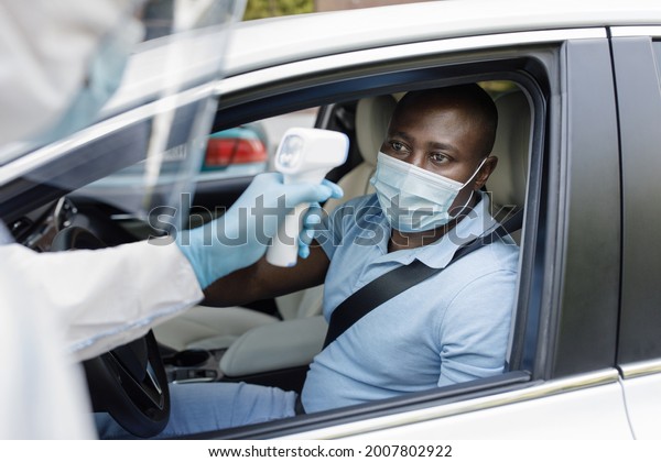 Black man driver wearing protective face\
mask, sitting inside white car, medical worker in protective suit\
checking african american guy body temperature with electronic\
thermometer before\
vaccination