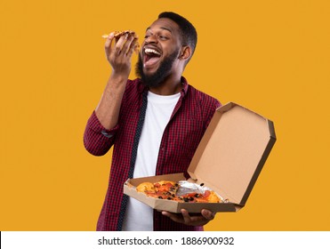 Black Man Eating Pizza Standing Holding Pizzeria Box Over Yellow Studio Background. African American Guy Enjoying Junk Food. Unhealthy Nutrition, Cheat Meal And Pizza Delivery Concept