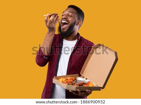 Black Man Eating Pizza Standing Holding Pizzeria Box Over Yellow Studio Background. African American Guy Enjoying Junk Food. Unhealthy Nutrition, Cheat Meal And Pizza Delivery Concept
