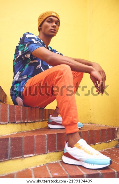 Black man, fashion and street style clothes on\
cool, trend or attitude model on steps by city wall background on\
building. Portrait of serious student sitting on stairs in urban\
punk clothing outdoor