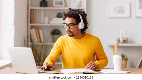 Black man freelancer in glasses and wireless headphones   working at  laptop while sitting at table and working remotely from home - Powered by Shutterstock