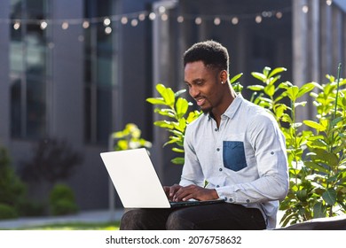 Black Man Freelancer Working Online Using Laptop Sitting On Bench Outside Office Modern Building In City Urban Park On Street. Happy African American Guy Student In Casual Outdoors Typing On Keyboard