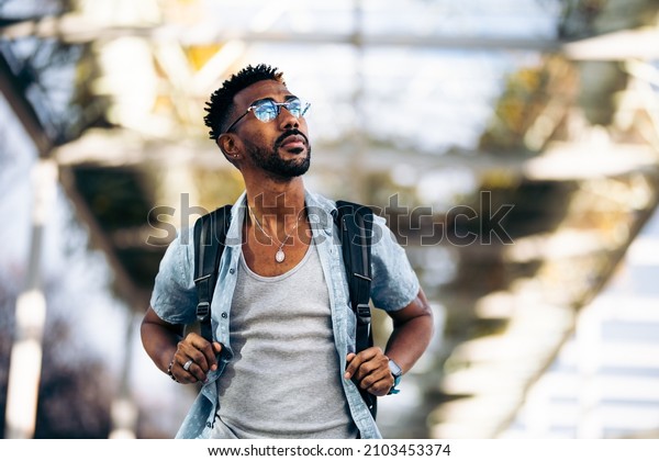 Black man with\
glasses and backpack walking down the street. Taking the handles of\
the backpack with his\
hands.