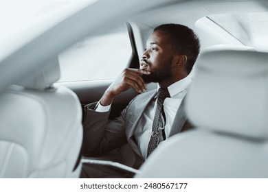 A black man in a grey suit with a white shirt and a dark tie sits in the backseat of a car looking out the window, deep in thought. His hand rests on his chin as he ponders. - Powered by Shutterstock