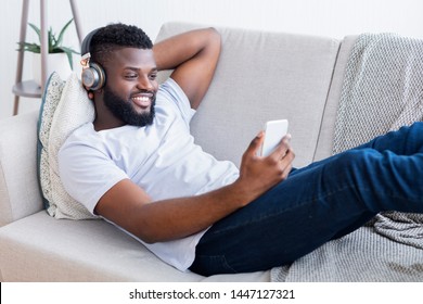 Black Man In Headphones Watching Movie On Smartphone, Lying On Couch At Home