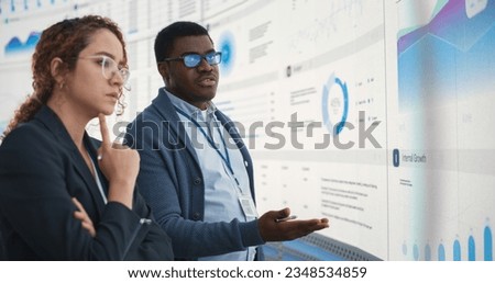 Black Man And Hispanic Woman Working On Business Strategy and Standing Infront of Big Digital Screen With Data. Male And Female Colleagues Discussing Plan For Profit Margins Enlargement In The Office.