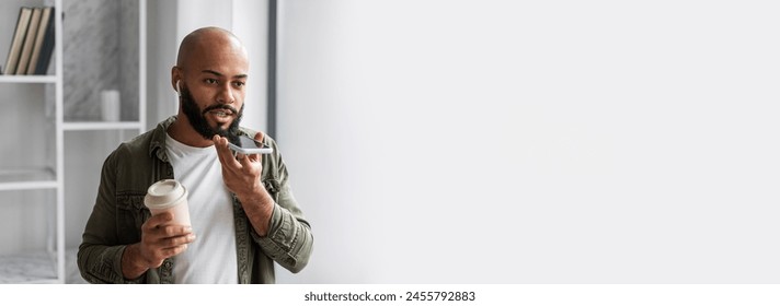 Black man holding a coffee and taking voice notes on a phone in a modern setting, with web-banner copy space - Powered by Shutterstock