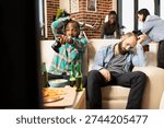 Black man holding wireless controller points at TV while seated next to upset caucasian friend after game loss. Diverse guys react to losing multiplayer video game tournament in cozy apartment.