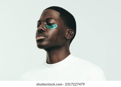 Black man indulging into some under eye care with a hydrogel patch. Standing in a studio with his yes closed, he absorbs the benefits of the beauty mask on his healthy and glowing melanin skin. - Powered by Shutterstock