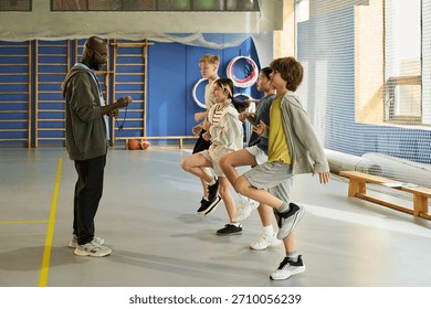Black man instructing group of multiethnic teenagers performing warm up exercises in gymnasium, teenagers lifting knees in coordinated movement, sports equipment visible in background