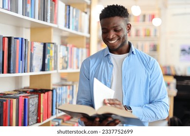Black man in a library or bookstore holding an open book and reading it while wearing a blue button-down shirt. The background consists of bookshelves filled with colorful books.