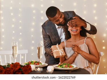 Black Man In Love Making Surprise To His Excited Woman On Valentine's Day, Man Closing Woman Eyes And Holding Gift Box, Restaurant Interior, Copy Space