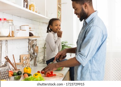 Black Man Making Food With His Sweet Daughter In Kitchen, Cutting Vegetables, Girl Eating Cookie And Smiling, Side View, Copy Space