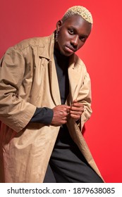 Black Man In Modern Trendy Beige Trench Coat Posing At Camera Isolated Over Red Background, Handsome Guy With White Dyed Hair Looking At Camera. People. Model, Fashion, Style Concept