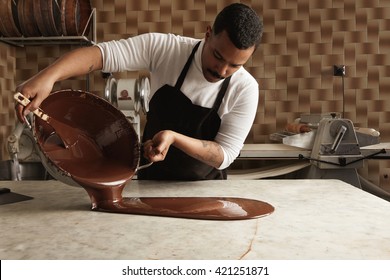 Black man professional chief pours tasty melted chocolate from one big steel pot to vintage marble table in his artisan rustic kitchen with industrial retro machines. Mirrored in chocolate on table.