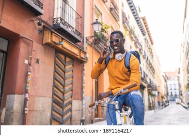 Black Man Riding Bike Having Phone Call Wearing Yellow Sweater Outdoors. African American Young Man Using Phone In The City. Lifestyle Concept.