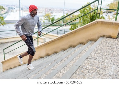 Black Man Running Upstairs Outdoors Listening To Music With White Headphones. Young Male Exercising With City Scape At The Background.