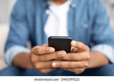 Black man seated on a couch, engrossed in his cell phone, scrolling and typing, cropped shot, closeup