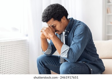 Black man is seated on a couch, with one hand pressed against his face in a contemplative gesture. He appears deep in thought, possibly experiencing emotions like stress, sadness, or exhaustion. - Powered by Shutterstock