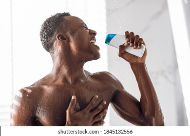 Black Man Singing In Shower Holding Shampoo Bottle Like Microphone Having Fun Bathing Standing In Modern Bathroom At Home. Hygiene And Male Bodycare Beauty Routine Concept.