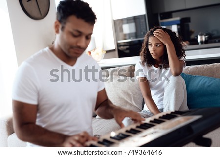 A black man sits in the living room of his apartment and plays a synthesizer. A girl sits next to him, covering her ears. A man is terribly playing and his girl is unhappy with this.