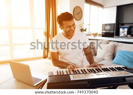 A black man sits in the living room of his apartment and plays a synthesizer. He composes music. Nearby is a gray laptop. A man likes to play music.