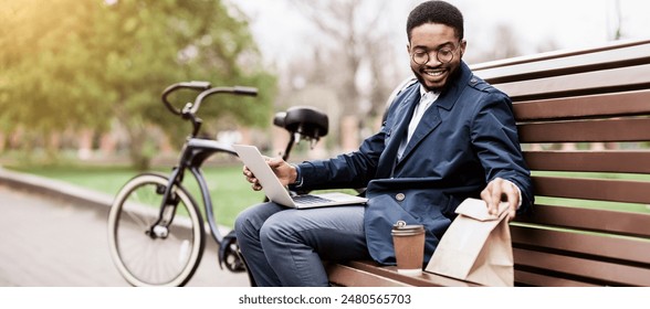 Black man sits on a park bench using his laptop, his bicycle nearby. The setting is calm and professional, reflecting a modern approach to work in a natural environment. - Powered by Shutterstock