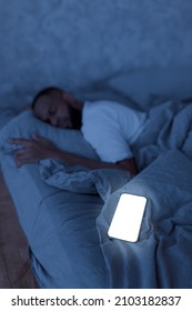 Black Man Sleeping, Resting In Bed In The Night At Home, Lying Near Smartphone, Vertical Portrait, Blurred Background, Selective Focus On Cell Phone With White Blank Screen For Mock Up Template