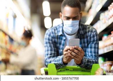 Black Man With Smartphone Shopping In Supermarket. African Guy Buying Food Wearing Face Mask And Using Grocery Shop App On Mobile Phone Walking Along Aisle And Store Shelves In Shop. Selective Focus