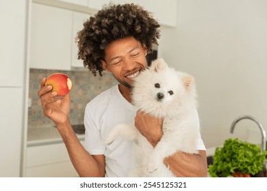 A black man smiles broadly in a bright kitchen, holding an apple in one hand and a fluffy white dog in the other. Fresh produce is visible on the counter, adding to the cheerful atmosphere. - Powered by Shutterstock