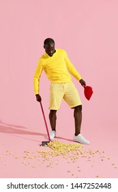 Black Man Sweeping Pop Corn On Floor With Broom On Pink Background. Full Length Portrait Of Male In Yellow Clothes Cleaning Popcorn Mess In Studio