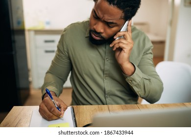 Black Man Taking Notes While Communicating Over Mobile Phone And Working At Home. 