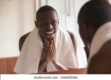 Black man towel on shoulders standing in bathroom look in mirror washes face touch skin after shaving or applying cream aftershave lotion feels satisfied, morning routine, personal hygiene, skincare