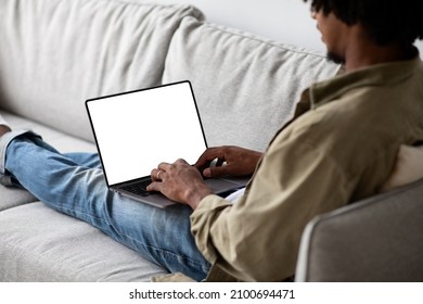 Black Man Using Laptop Computer With Blank White Screen While Sitting On Couch At Home, African American Guy Working Online Or Browsing Internet Website, Over Shoulder View, Mockup With Copy Space