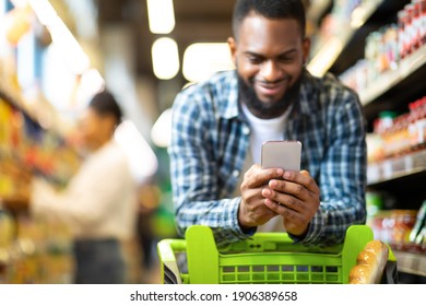 Black Man Using Mobile Coupon App For Groceries Shopping Buying Food In Supermarket, Standing With Cart In Hypermarket. African Guy Using Smartphone Purchasing Grocery In Shop. Selective Focus