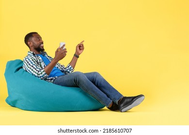 Black Man Using Mobile Phone Pointing Finger Aside Sitting In Beanbag Chair Over Yellow Studio Background. Guy Advertising Great Application Via Cellphone. Side View Shot - Shutterstock ID 2185610707