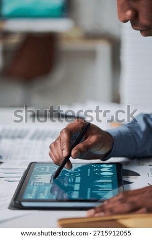 Black man using stylus interacting with digital tablet displaying data charts and analytics, close up of hand and device on desk, adult engaged in technology related task