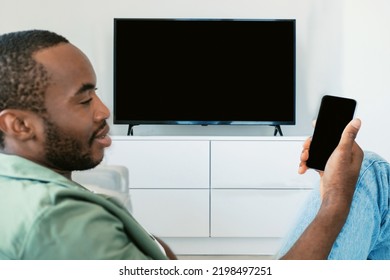 Black Man Watching Television With Blank Screen And Using Smart TV Remote Control App On Smartphone, Mockup. Select Movie Stream, Change Channel Or Settings In The Menu