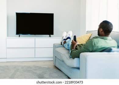 Black Man Watching TV With Blank Screen, Holding Remote Control And Switching Channels On Flatscreen Plasma, Sitting On Sofa At Home, Spending Weekend Indoors. Mockup