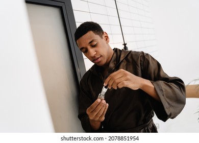 Black Man Wearing Bathrobe Applying Face Serum At Morning In Bathroom