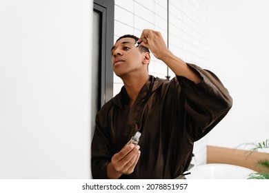 Black Man Wearing Bathrobe Applying Face Serum At Morning In Bathroom